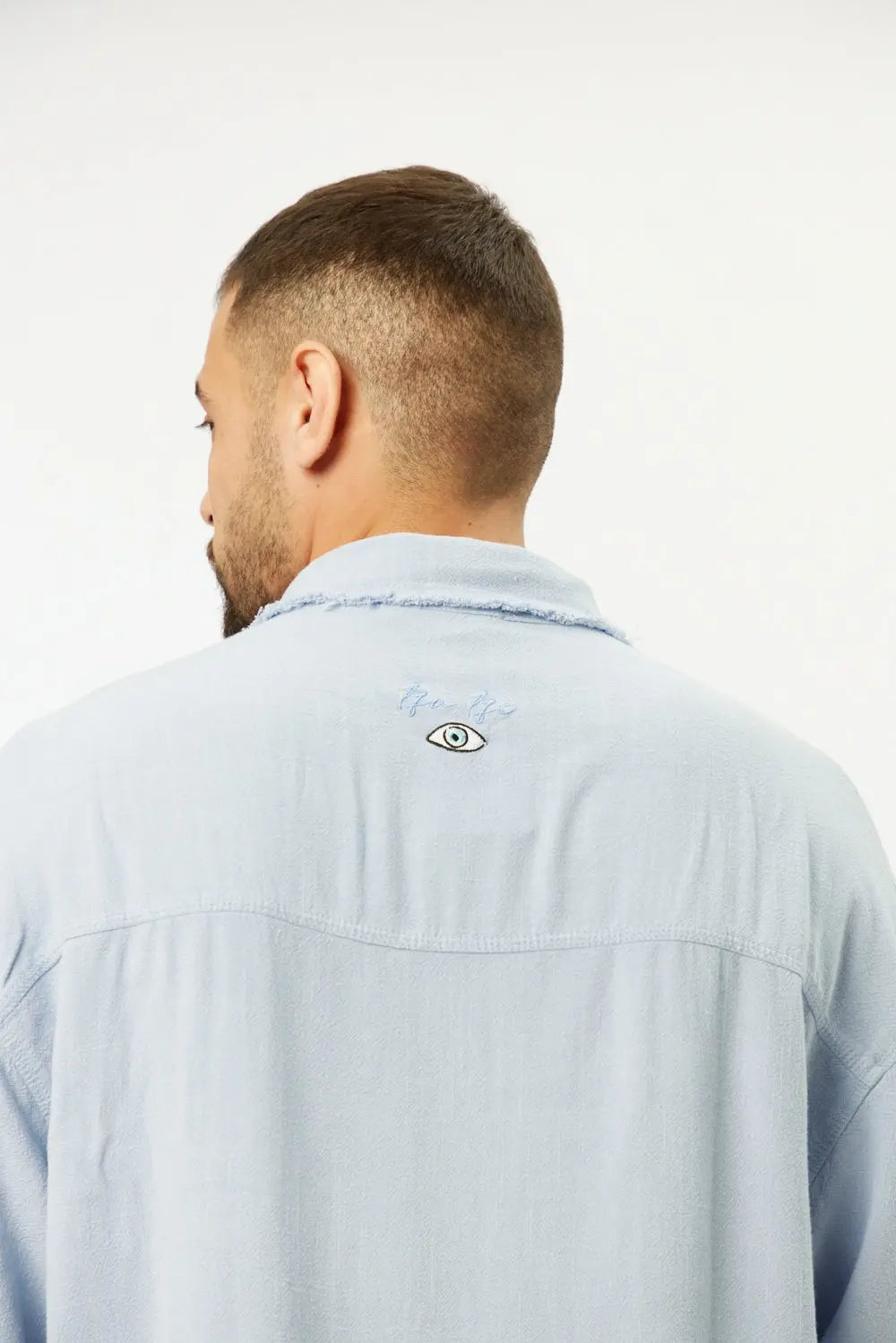 Back of a man wearing a sky blue Soft Linen Short Sleeve Button-Up Shirt, showing an embroidered eye graphic on the upper back