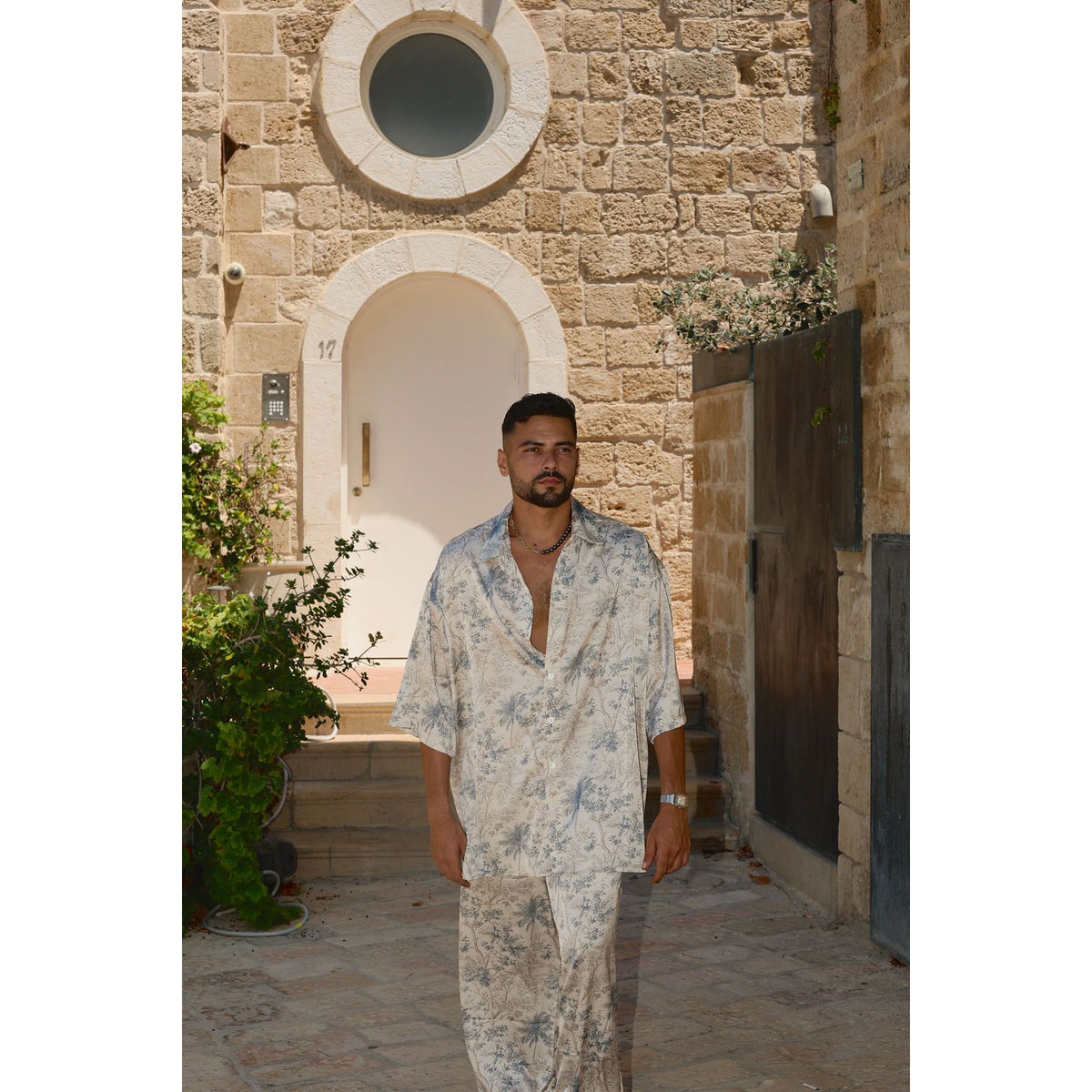 A man wearing a light blue and white tropical floral print lounge set stands in front of a stone building with a white arched doorway.