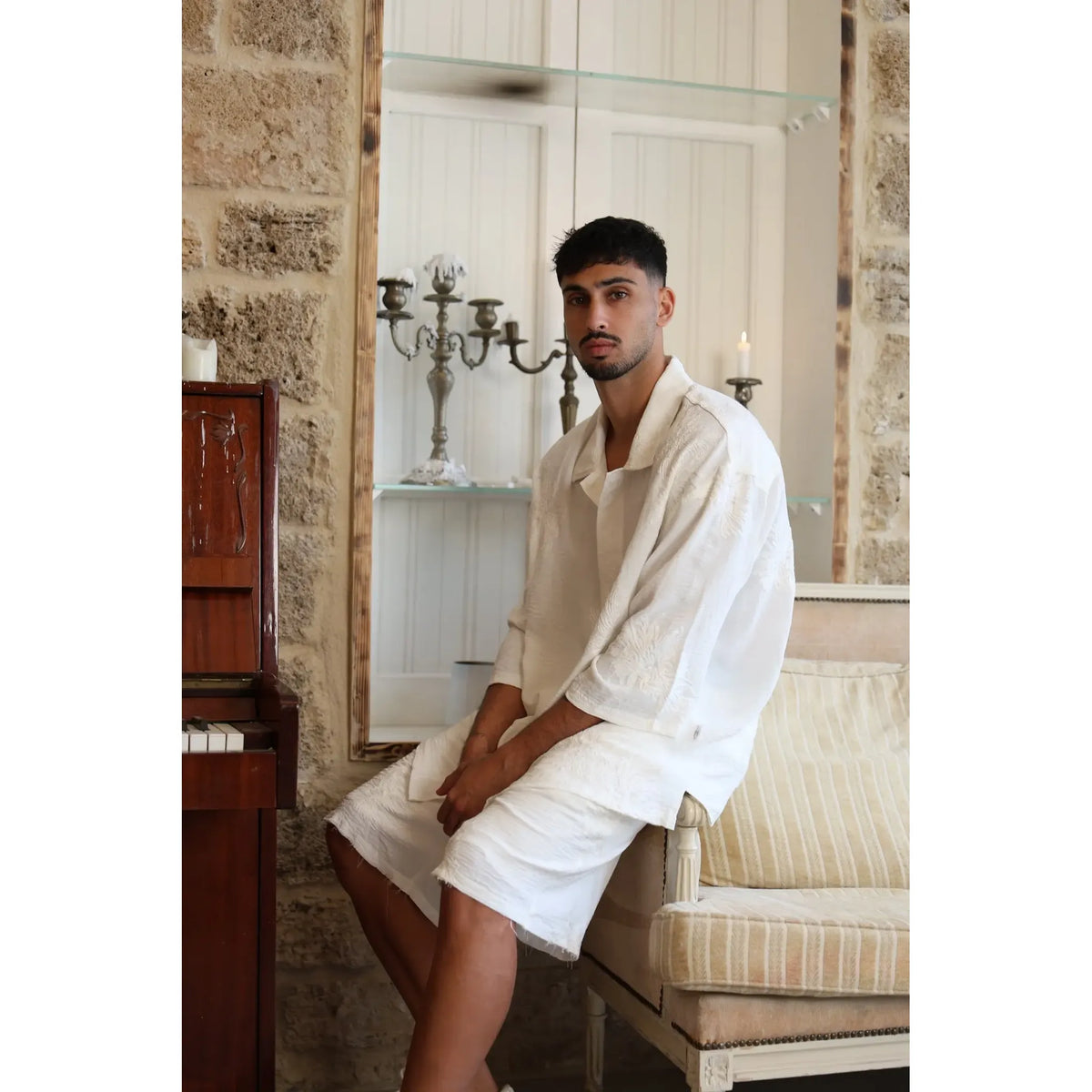 Man wearing a white SPARKS 3/4 Sleeve Button-Up Shirt and white shorts, sitting on a sofa next to a piano