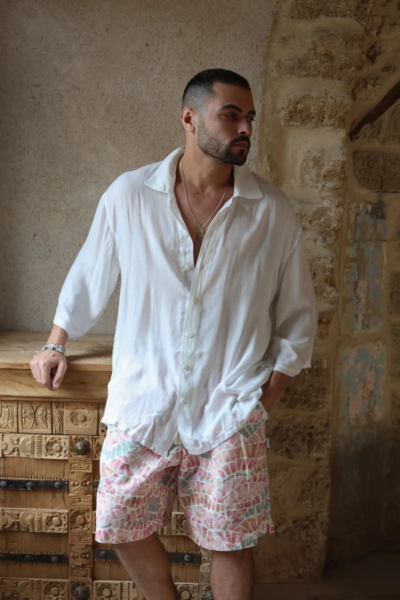 Man wearing a white VENICE Button-Up Shirt and patterned shorts, leaning on a carved wooden cabinet
