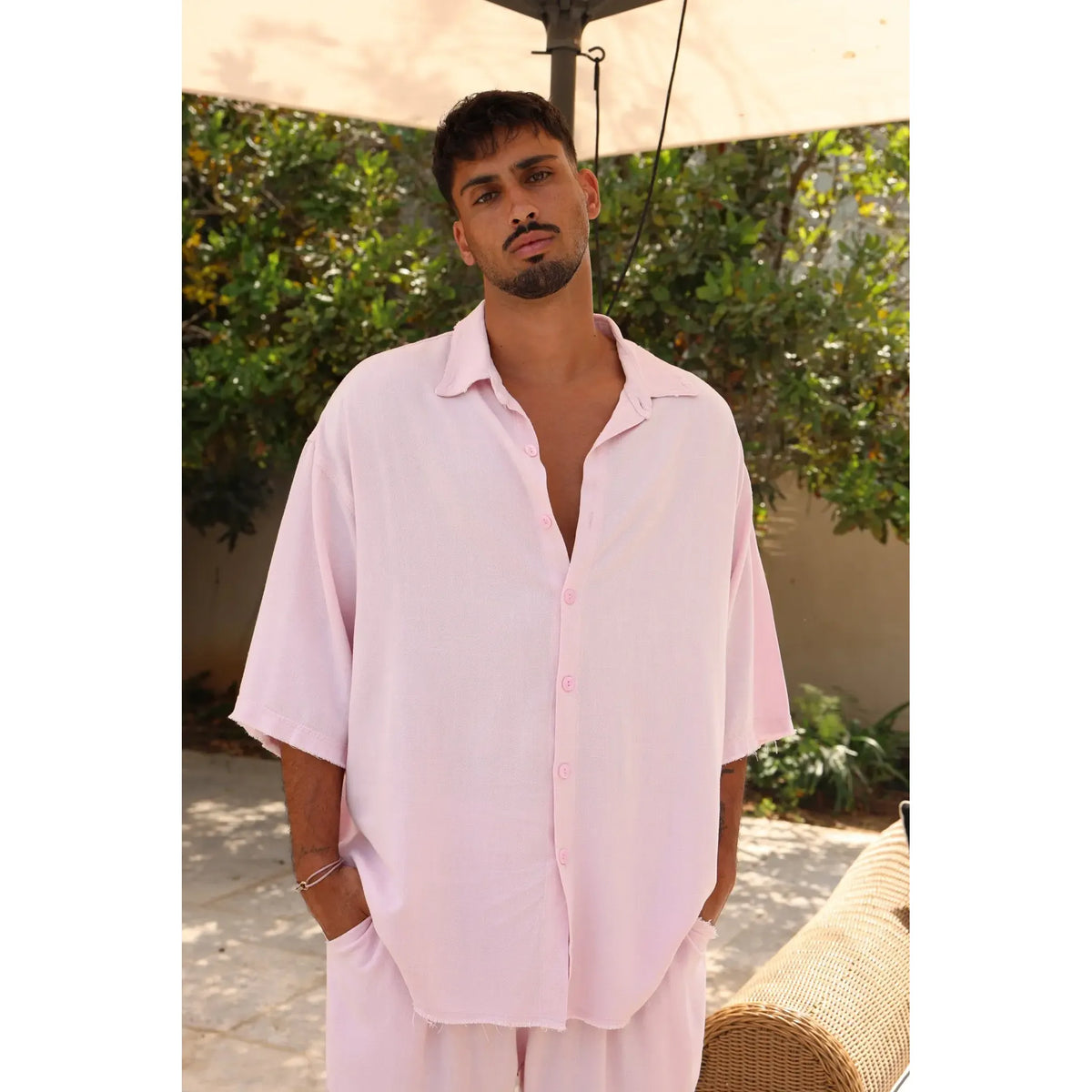 Man wearing a light pink shirt and pants standing outdoors with greenery in the background