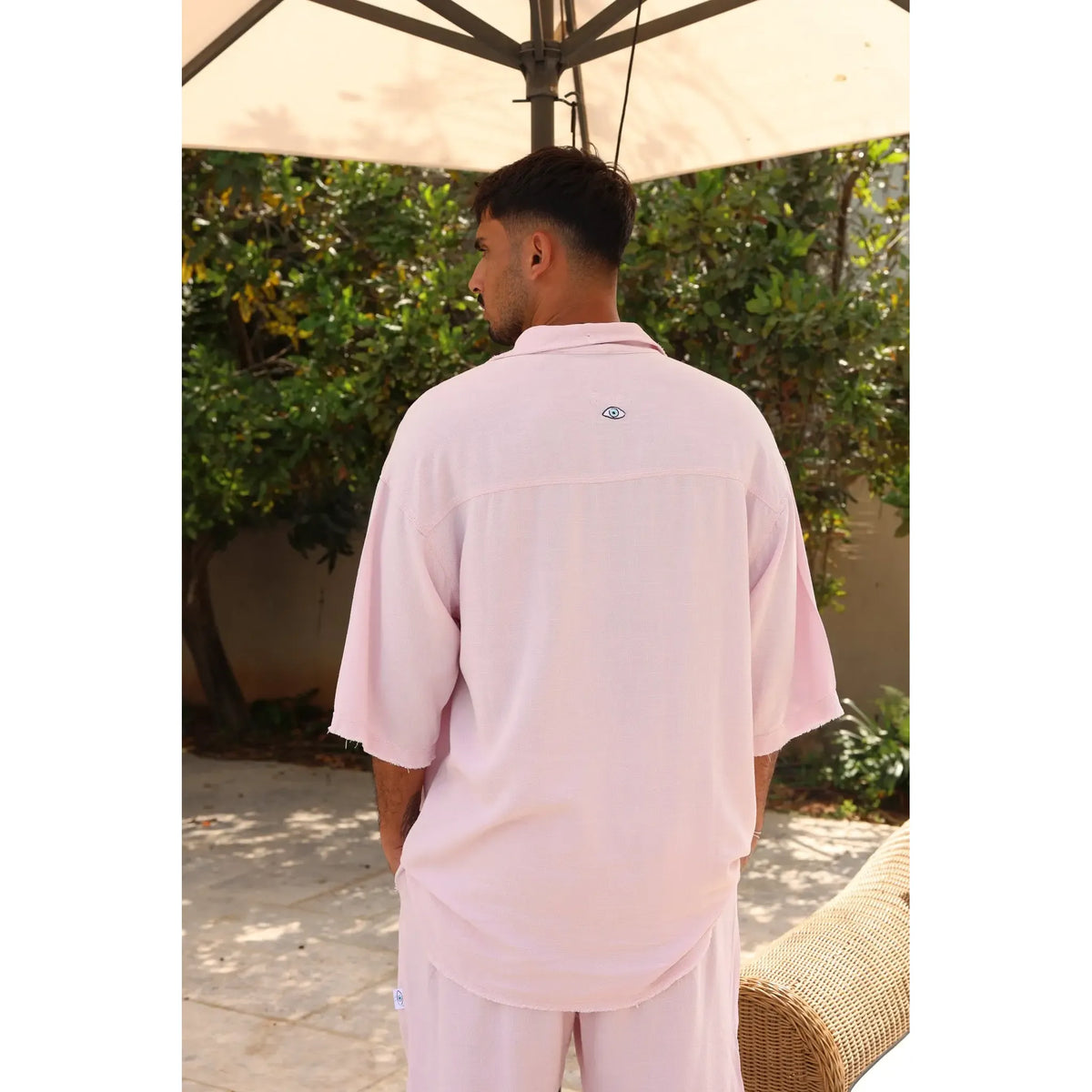 Man from behind wearing a Soft Linen Short Sleeve Button-Up Shirt in Baby Pink, standing outdoors under an umbrella