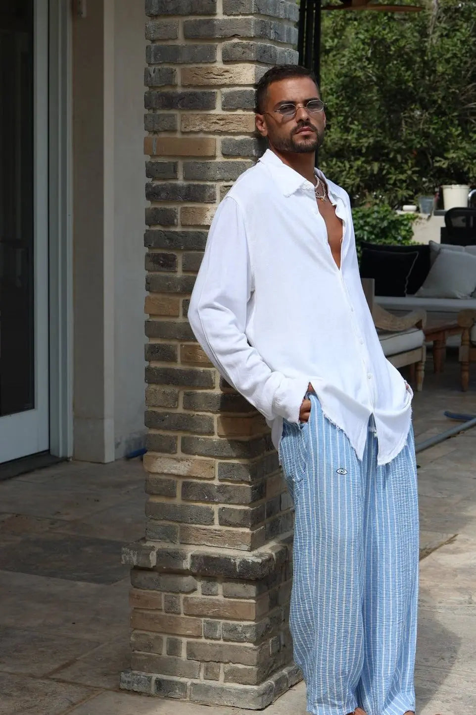 Man wearing Pure Oversized Linen Pants in Sky Blue Stripes and a white shirt, leaning against a brick pillar
