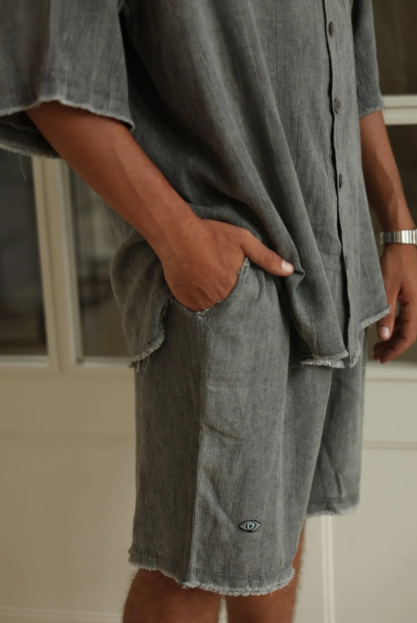 Man wearing Soft Linen Bermuda Shorts in Steel Grey, with hand in pocket, showing the eye logo