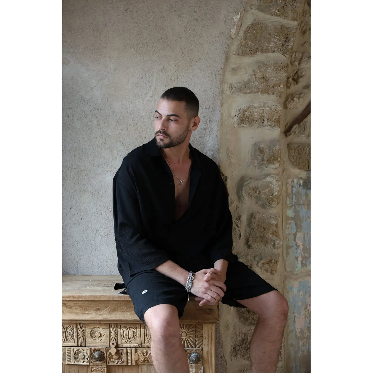 Man wearing a black SPARKS 3/4 Sleeve Button-Up Shirt and black shorts, sitting on a wooden chest