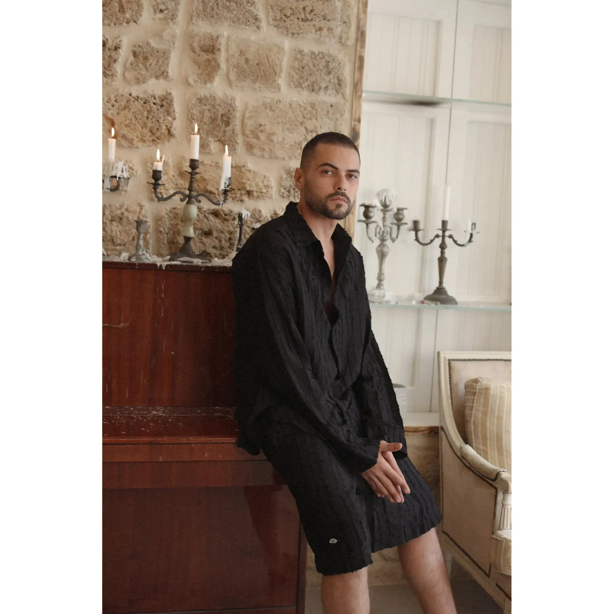 Man wearing a black RIPS Long Sleeve Button-Up Shirt and black shorts, leaning on a wooden piano with lit candles in the background
