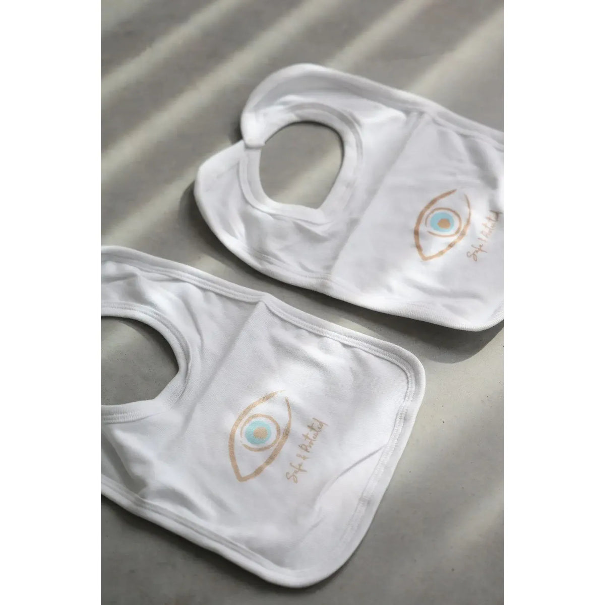 Two white Newborn Cotton Bibs with an eye graphic and Safe & Protected text