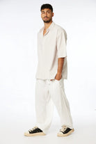 Man wearing a Striped Linen Short Sleeve Button-Up Shirt in Stone, white linen pants, and black and white sneakers