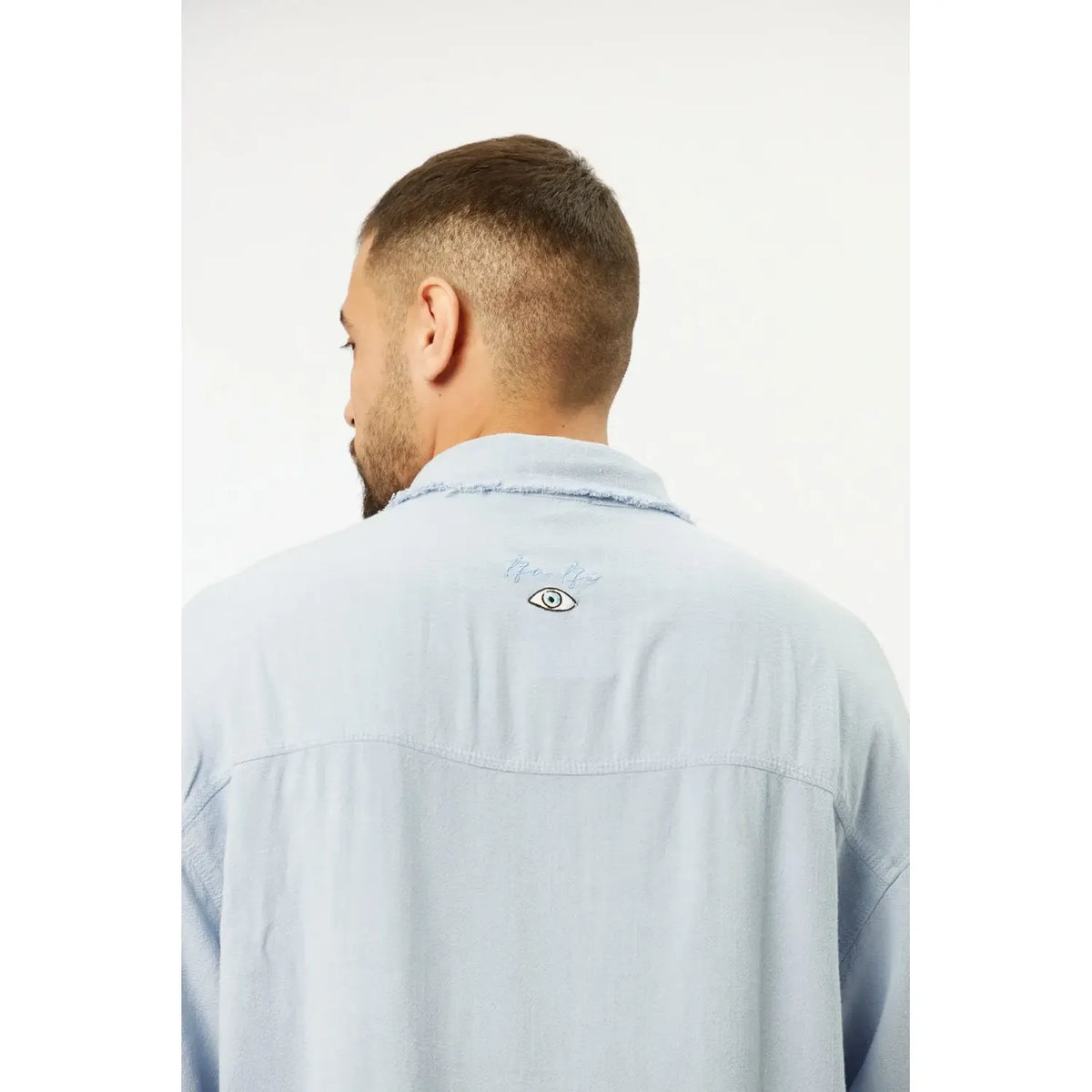 Back of a man wearing a sky blue Soft Linen Short Sleeve Button-Up Shirt, showing an embroidered eye graphic on the upper back