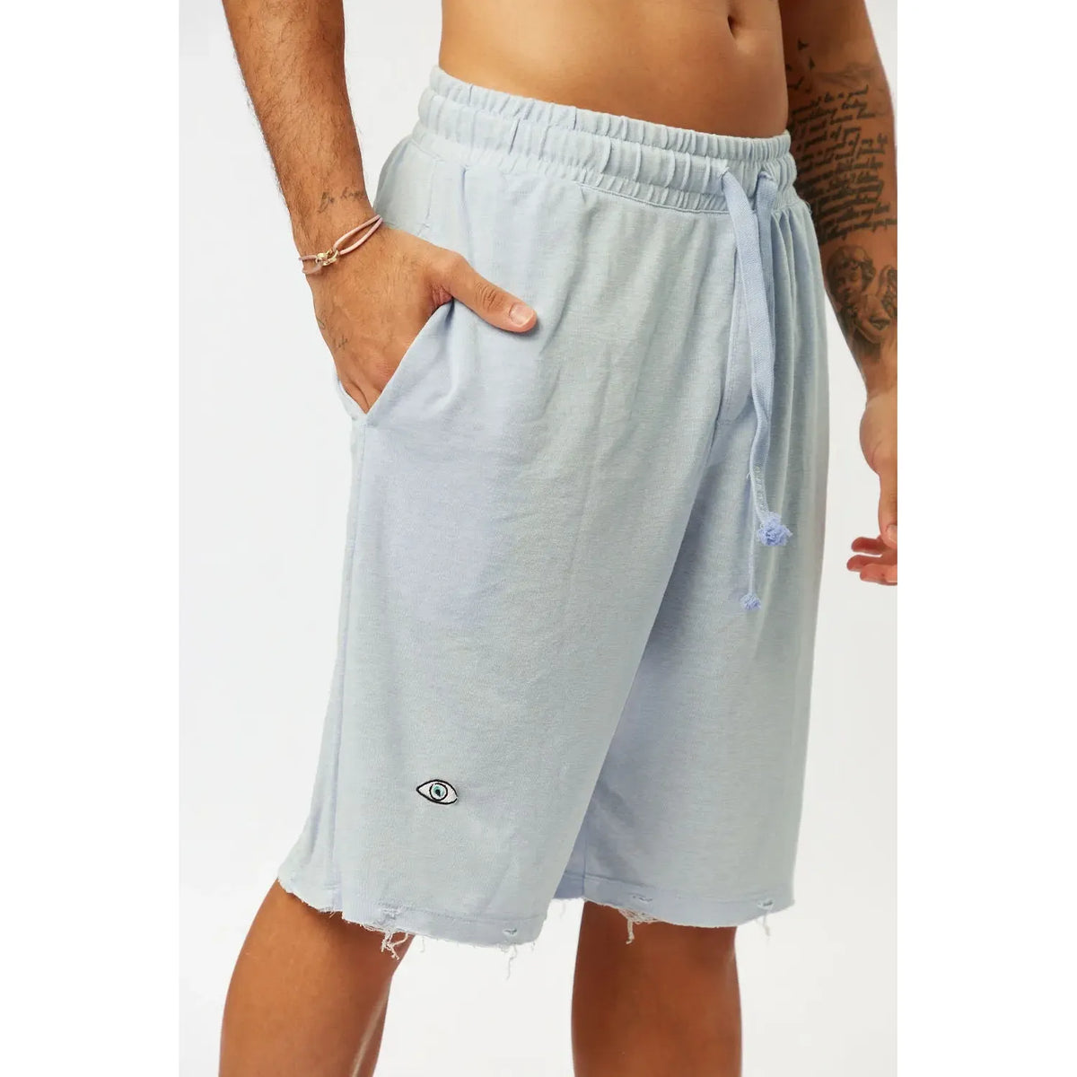 BASE Shorts in light blue with an eye logo and distressed hem, worn by a person with a hand in the pocket