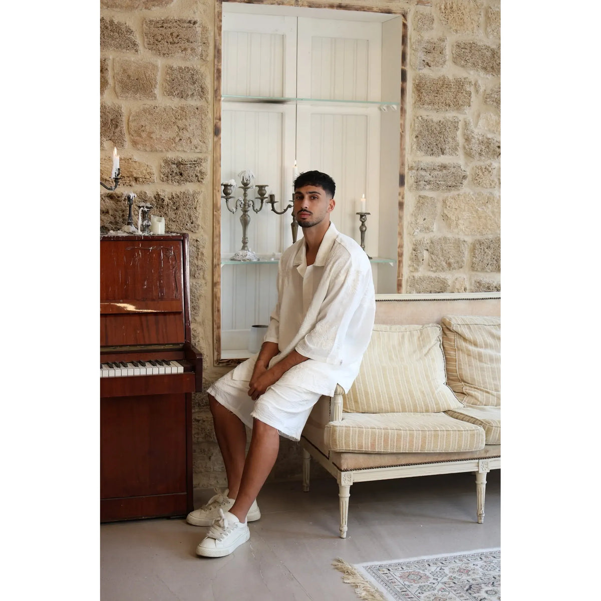 Man wearing a white SPARKS 3/4 Sleeve Button-Up Shirt and white shorts, sitting on a sofa next to a piano