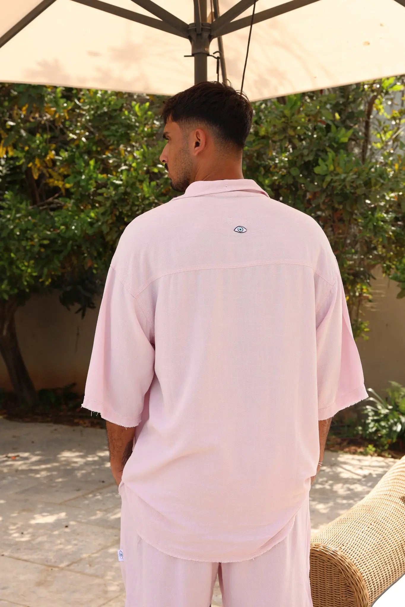 Man from behind wearing a Soft Linen Set in Baby Pink, short sleeve shirt and Bermuda shorts, with an eye logo on the back