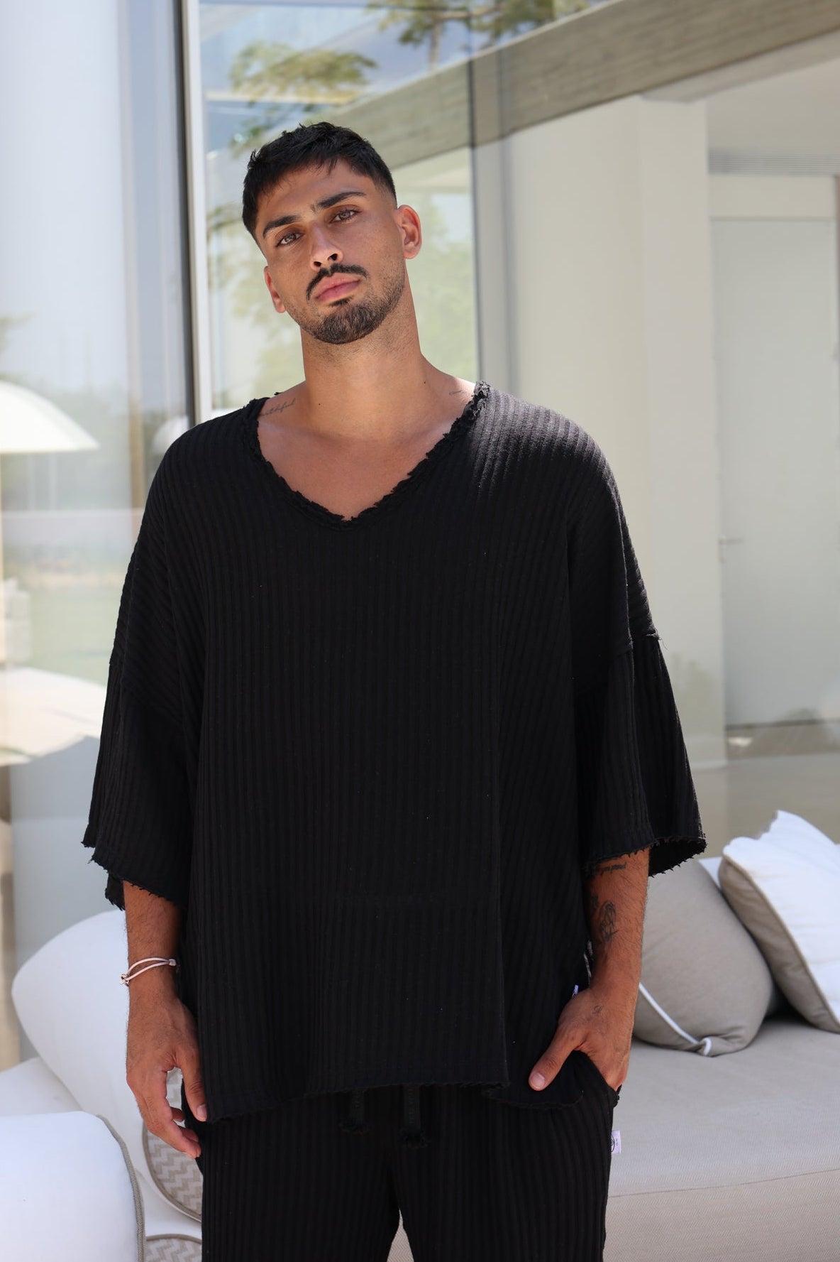 A man wearing a black ribbed V-neck tunic top with wide, tiered sleeves and matching black pants, standing in a modern, sunlit room with a white sofa in the background.