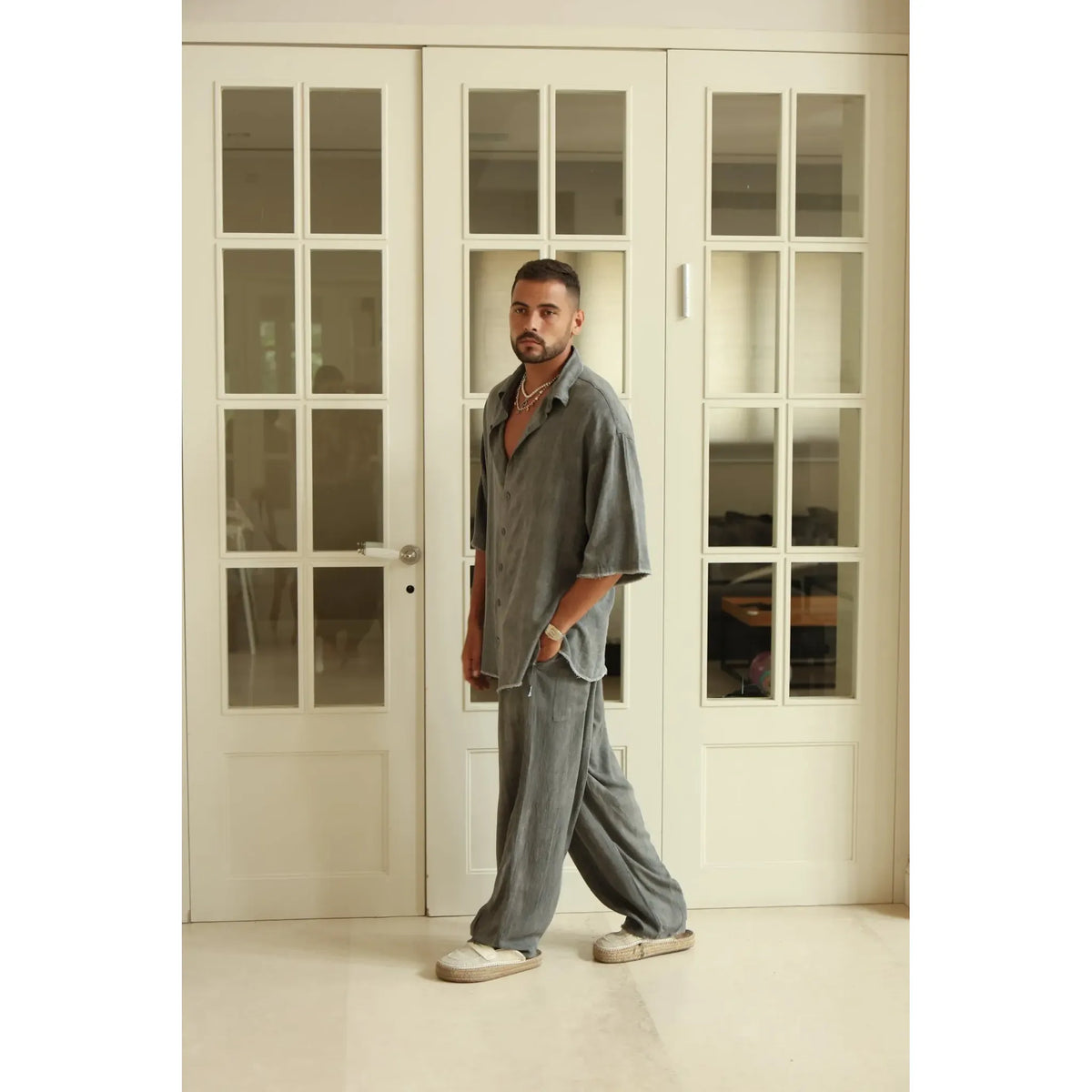 Man wearing Pure Grey Oversized Soft Linen Pants and matching shirt, standing in front of white glass-paned doors