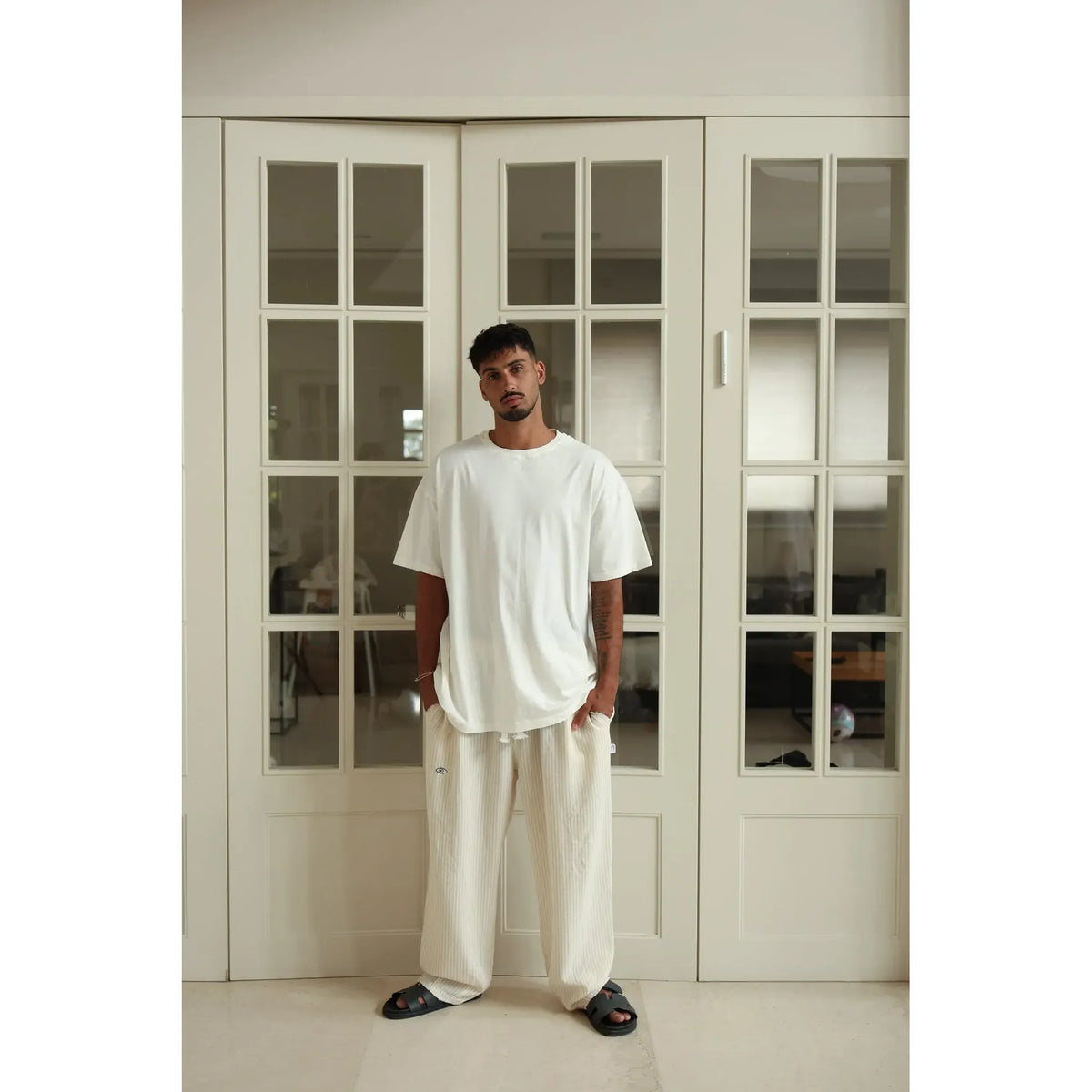 Man wearing a white t-shirt and pants standing in a room with large glass doors.