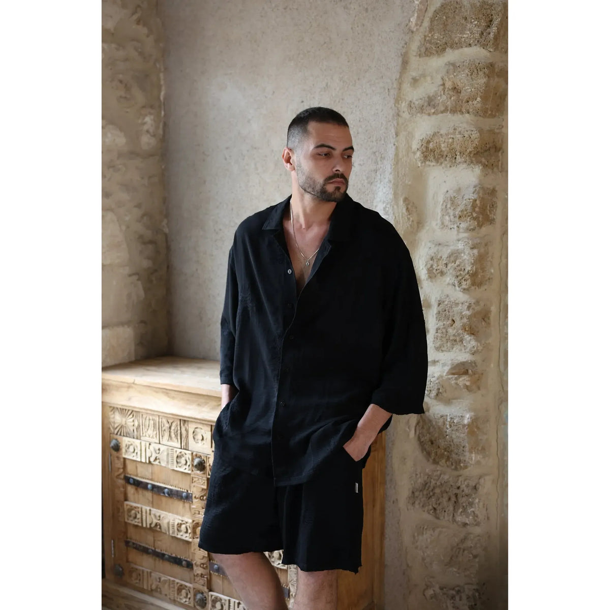 Man wearing a black SPARKS 3/4 Sleeve Button-Up Shirt and black shorts, standing next to a carved wooden cabinet