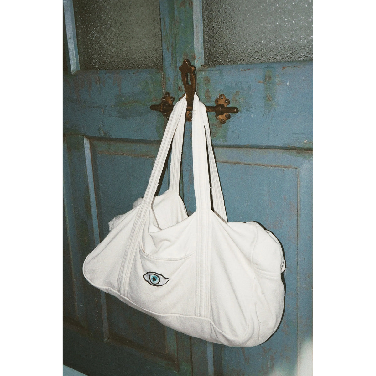White tote bag with a blue eye design hanging on a rustic wooden door.