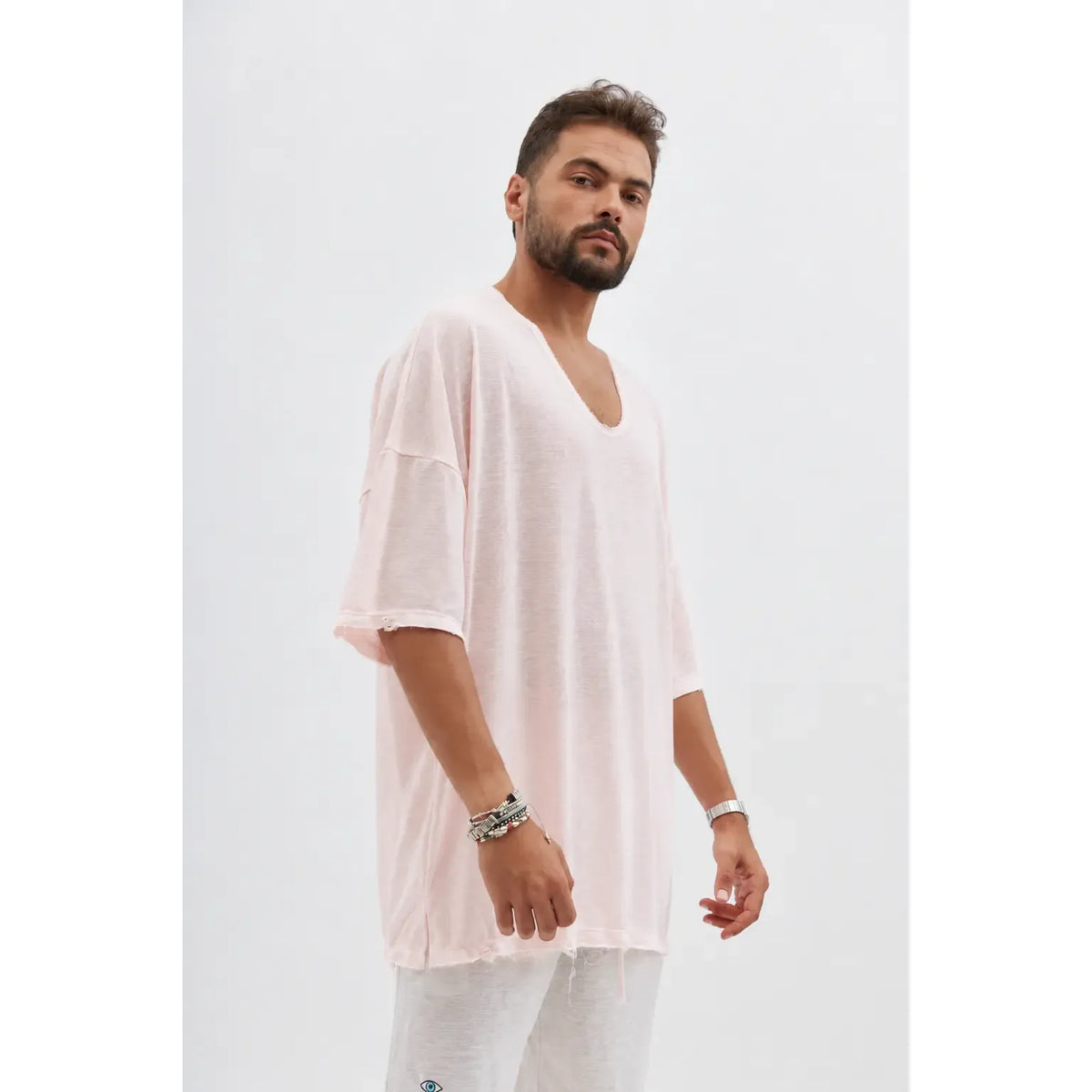 Man wearing a Dor Oversized Shirt in baby pink with a deep V-neck and distressed edges, paired with light grey trousers