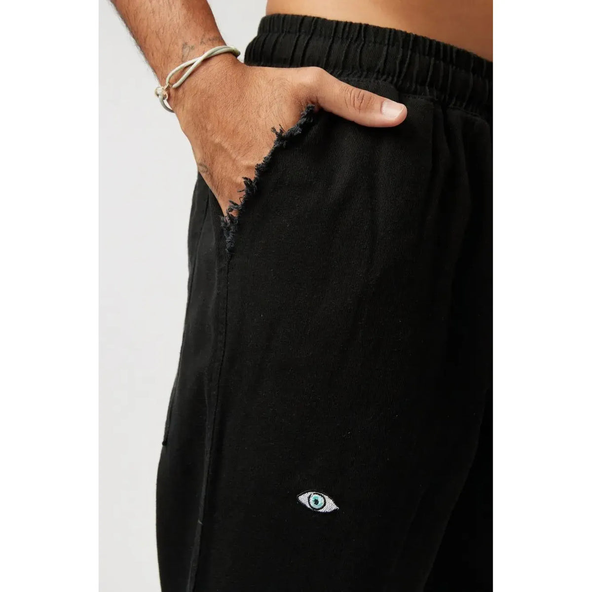 Close up of DC Elastic Waist Pants with Elastic Hem in black, showing a hand in a distressed pocket and an embroidered eye graphic
