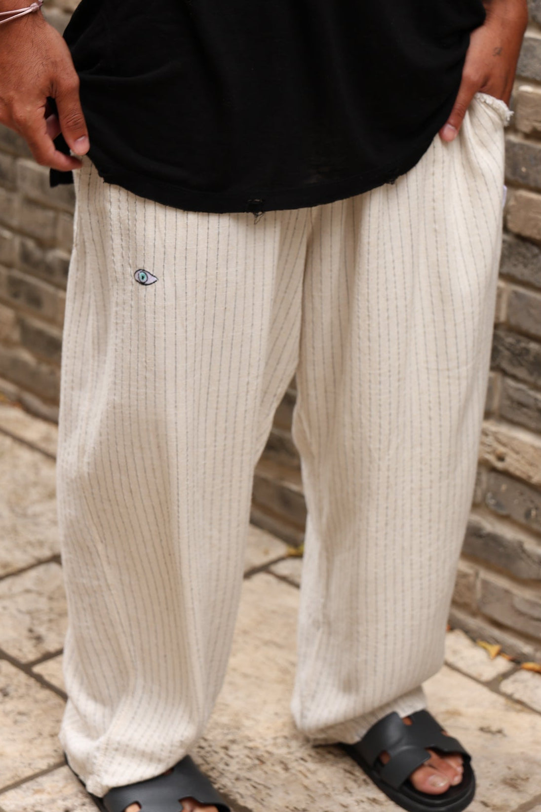 Person wearing beige pants with a small logo, standing on a stone path with a brick wall background.