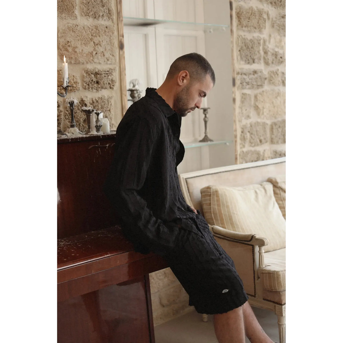 Man wearing a black RIPS Long Sleeve Button-Up Shirt and black shorts, leaning on a wooden piano