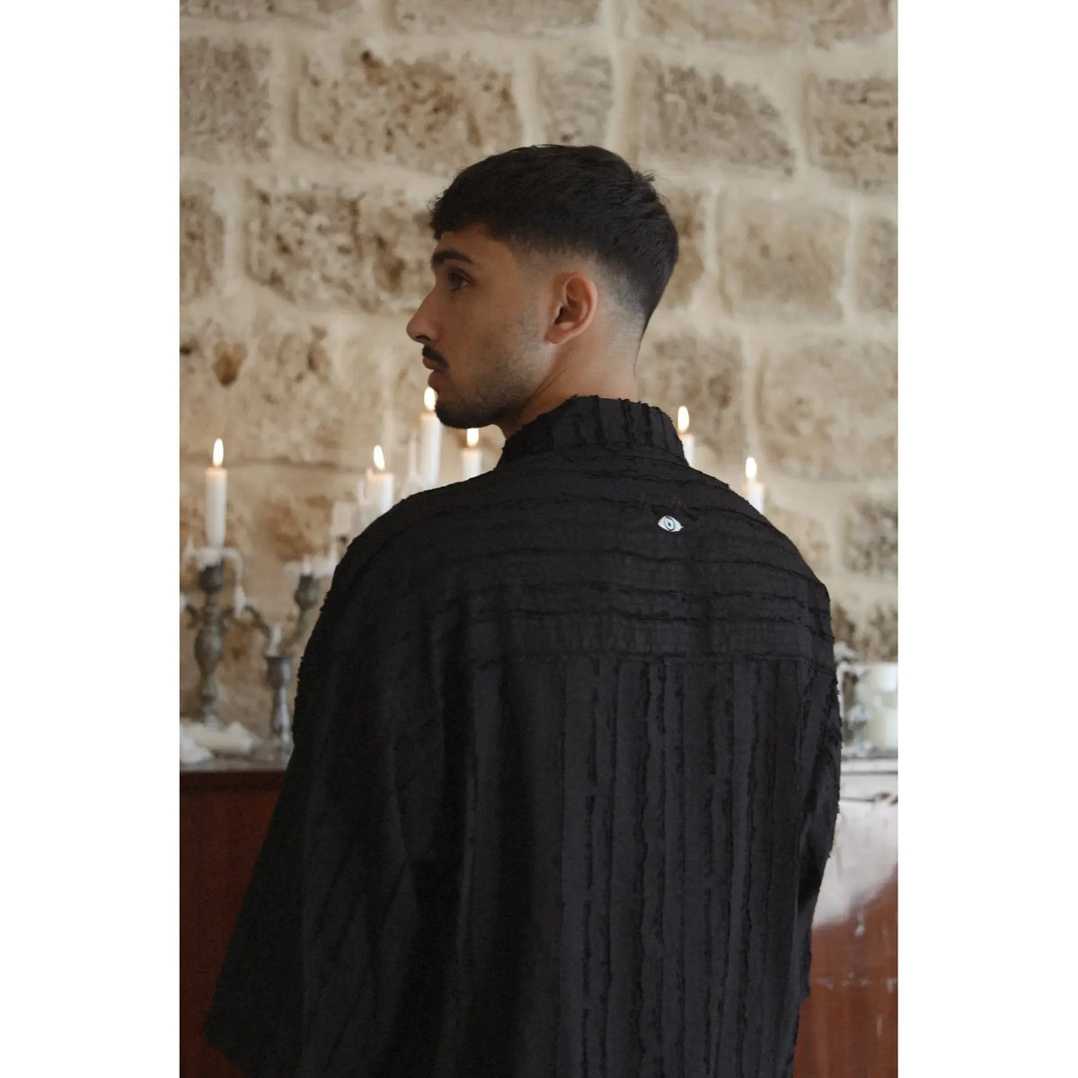 Man wearing a RIPS Short Sleeve Button-Up Shirt in black, facing away from the camera, looking left. A stone wall and lit candles are in the background