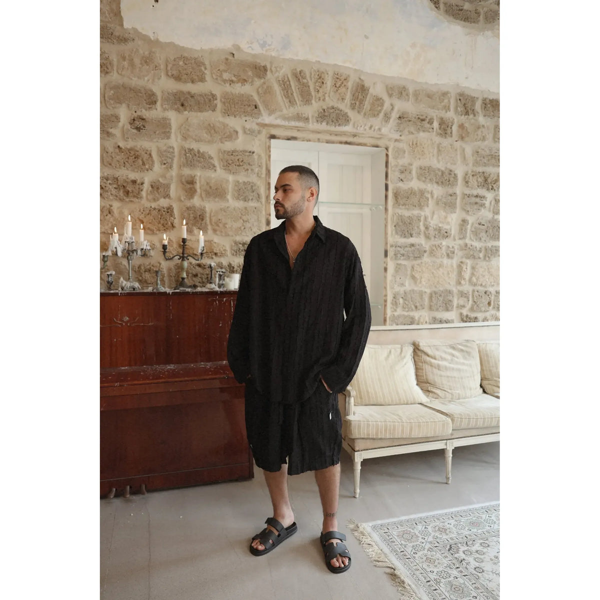 Man wearing a black RIPS Long Sleeve Button-Up Shirt and black shorts, standing indoors with a stone wall background