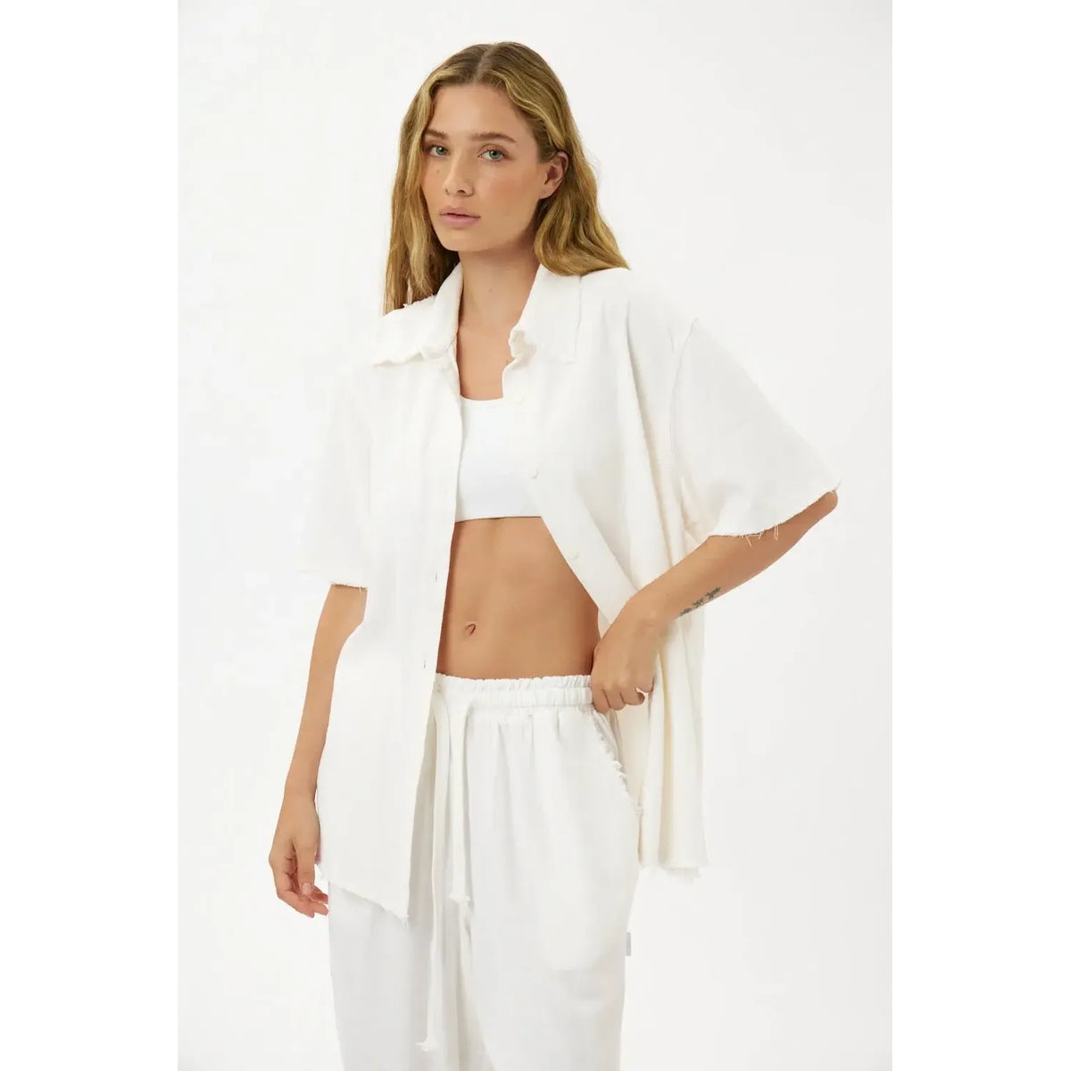 Woman wearing a white Soft Linen Short Sleeve Button-Up Shirt, unbuttoned over a white bralette, and matching white linen pants
