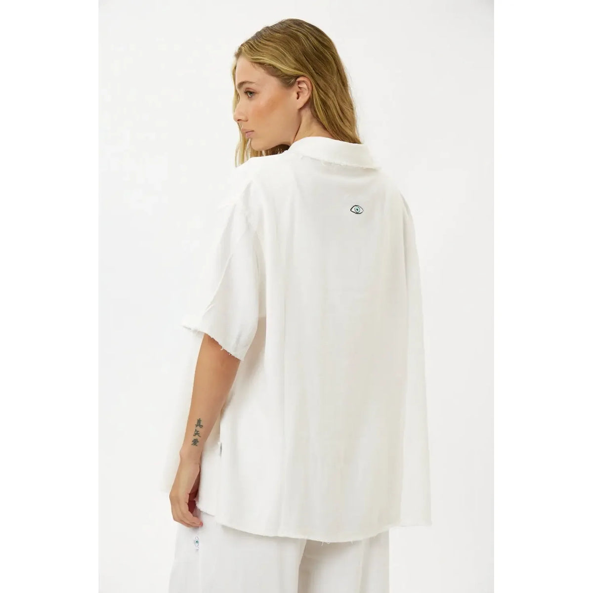 Woman wearing a white Soft Linen Short Sleeve Button-Up Shirt, seen from the back. The shirt has a small eye graphic on the upper back