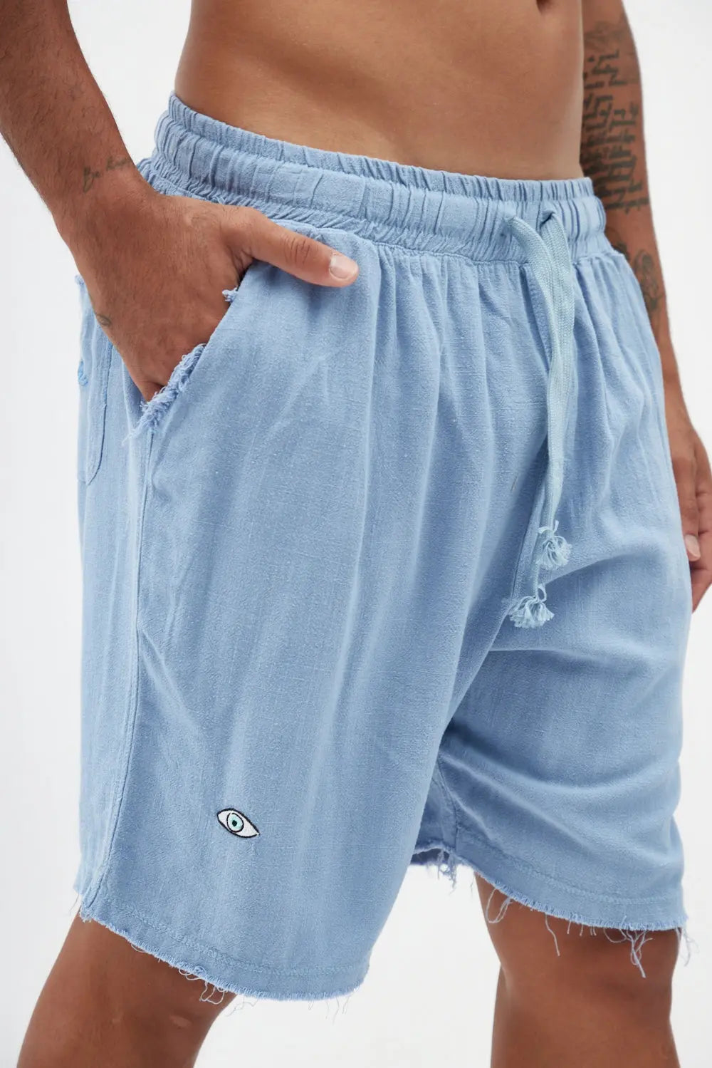 Close up of the Soft Linen Set Sky Blue Bermuda Shorts with a hand in the pocket and an eye logo on the left leg