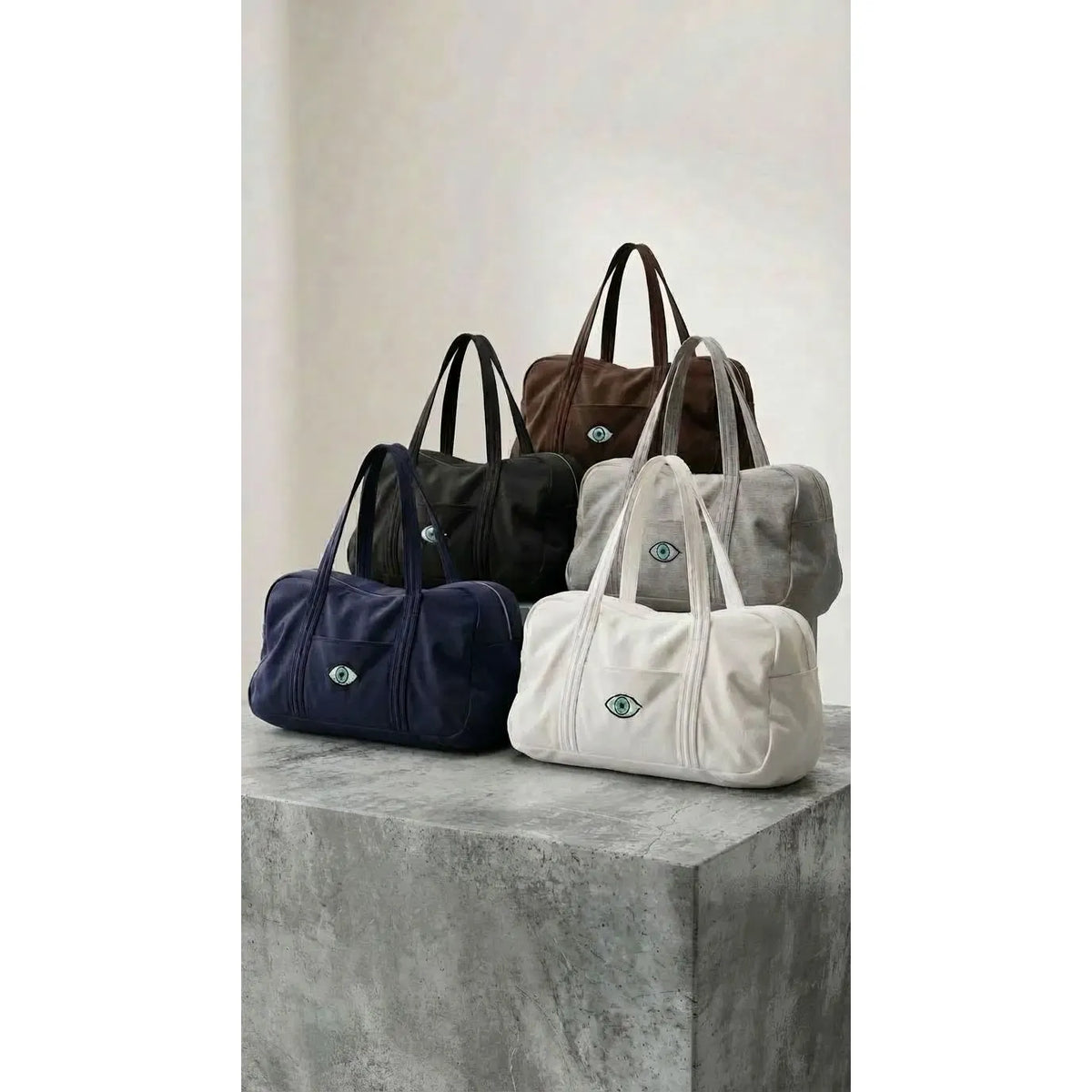Five Cotton Bags in black, brown, grey, navy, and white, each with an eye design, arranged on a concrete surface
