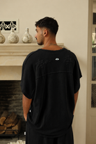 Man wearing a black t-shirt with a logo, standing in a room with decorative elements.
